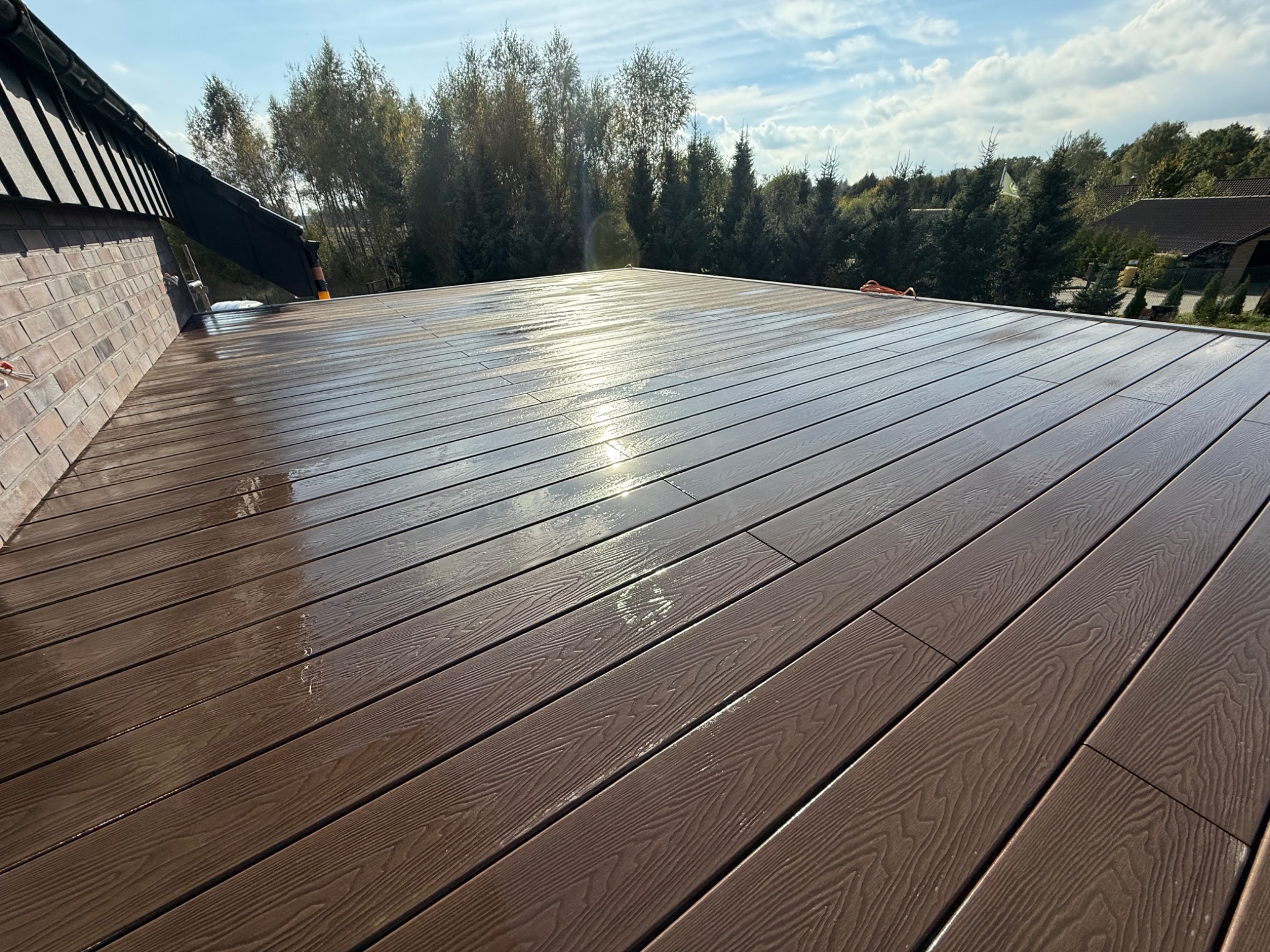 Błyszczący, brązowy taras kompozytowy po deszczu, z widoczną fakturą drewna i odbiciem słońca, na tle zieleni i błękitnego nieba z chmurami.