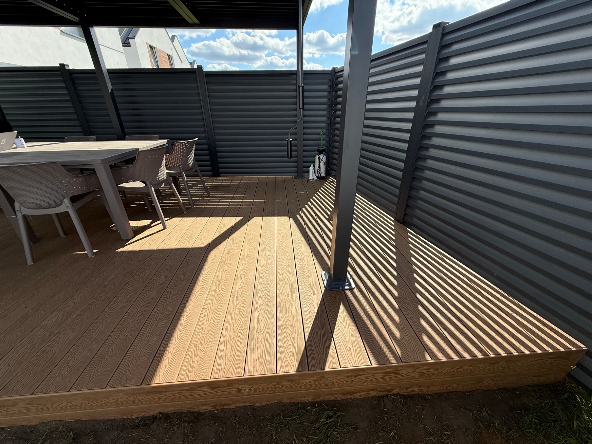 Jasny taras kompozytowy z szarą pergolą i ścianą z paneli. Na tarasie stół z krzesłami. Słoneczne cienie na desce. Nowoczesny design.