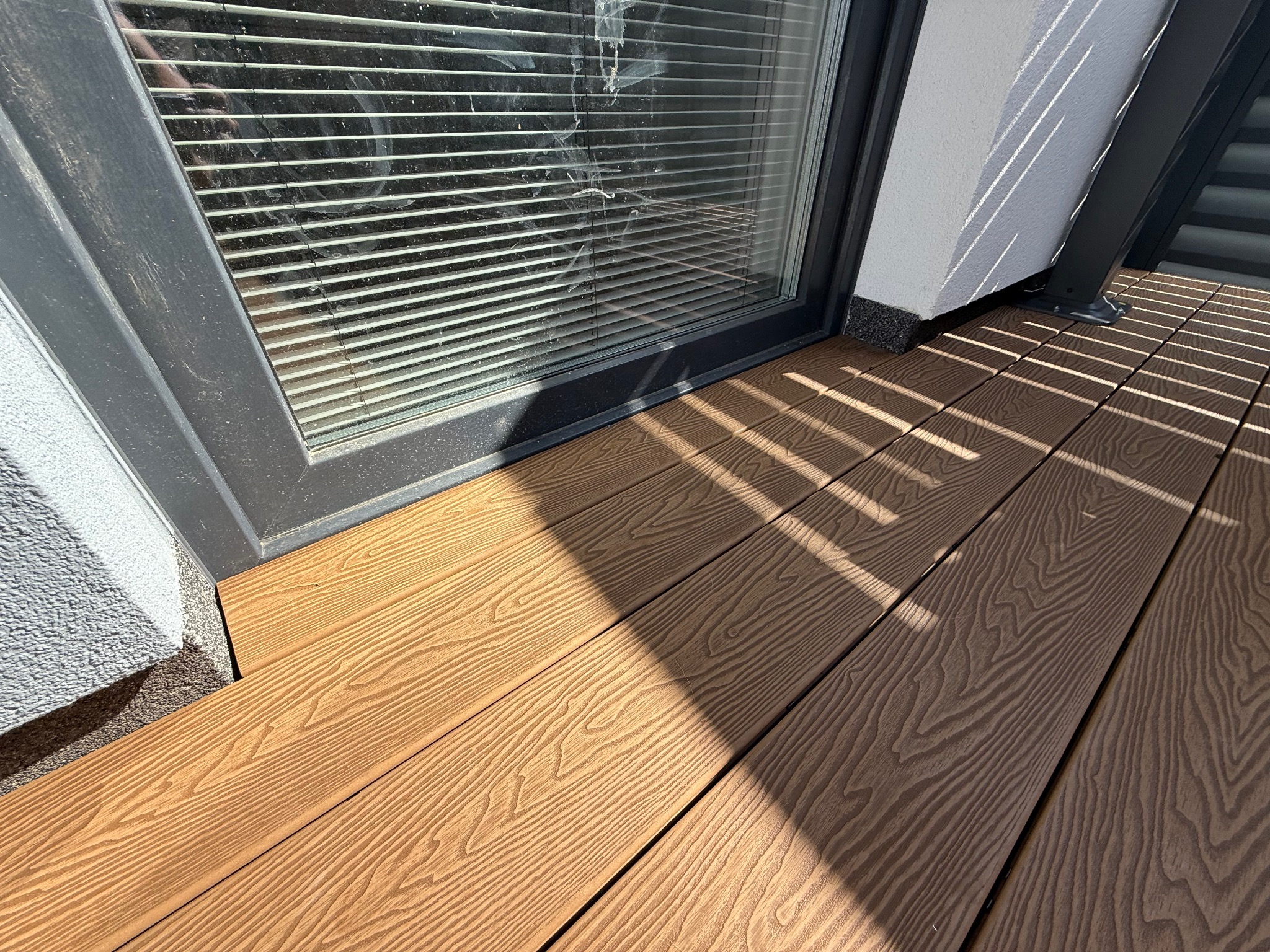 Kompozytowa deska tarasowa w odcieniach brązu, ułożona przy oknie z żaluzjami, z widocznymi promieniami słońca tworzącymi geometryczne cienie. Detal wykończenia tarasu.