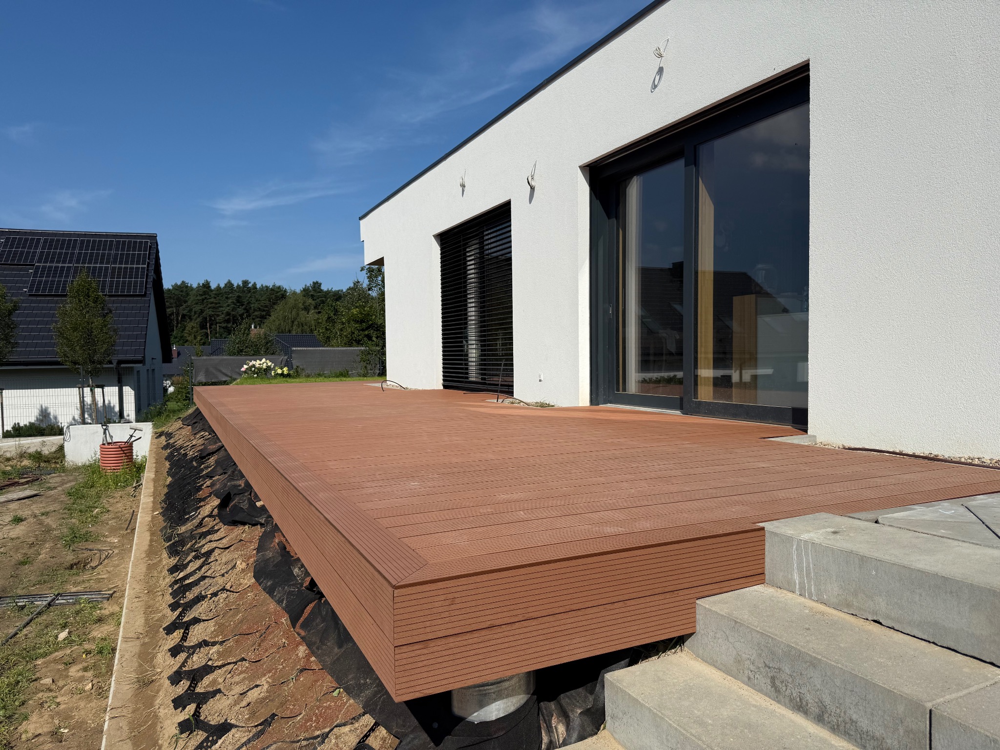 Nowoczesny taras z deski kompozytowej w kolorze brązu, przylegający do białego budynku z dużymi oknami. Widoczne schody wejściowe i fragment ogrodu w tle.