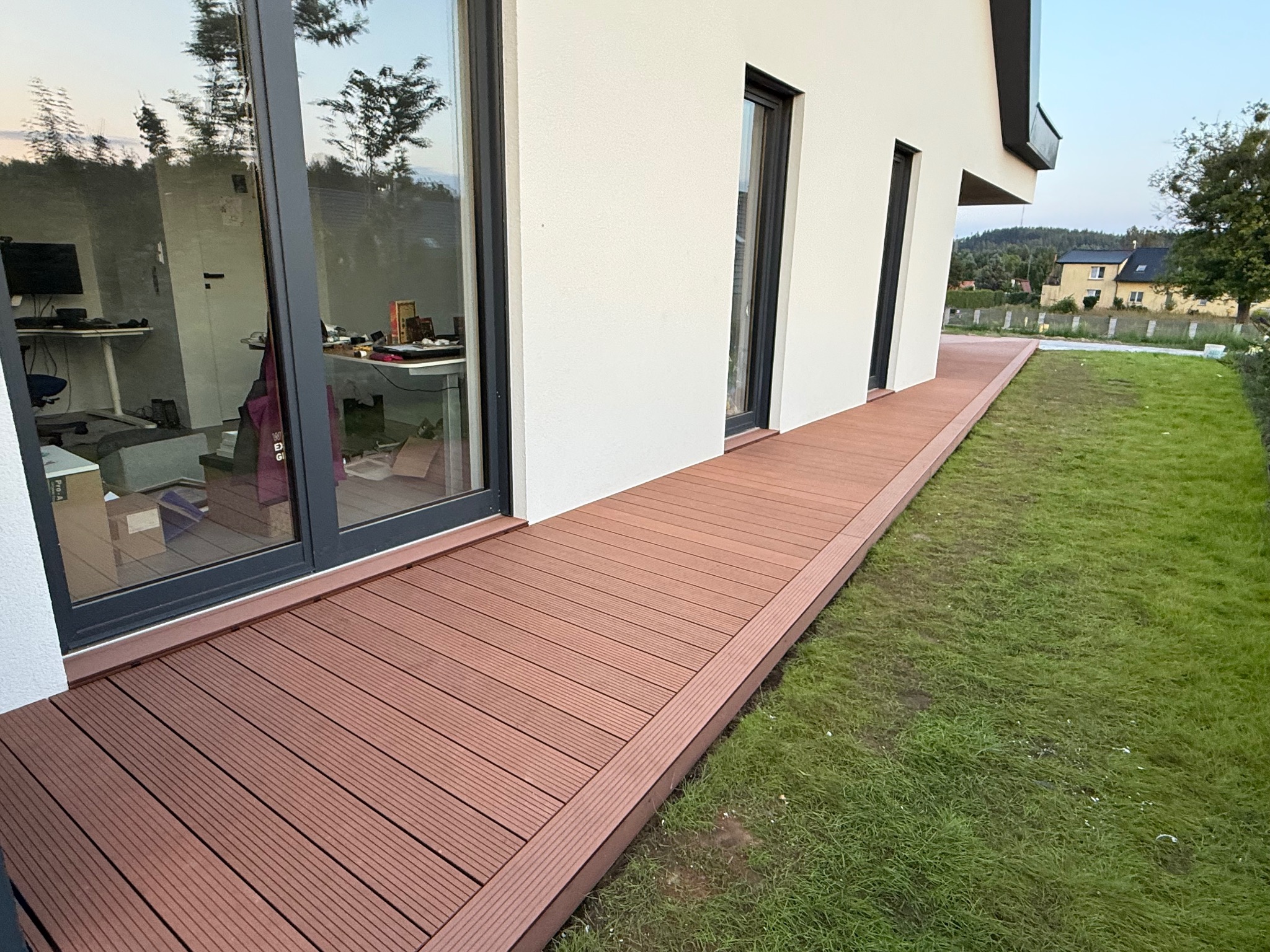 Nowoczesny taras z deski kompozytowej w kolorze brązu, przylegający do białego budynku z dużymi oknami. Widok na zadbany trawnik i zabudowę w tle.