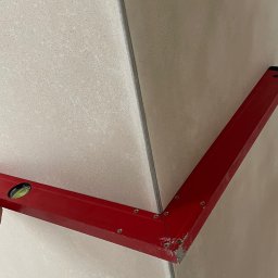 Portal-tech - Poziomica kątowa na jasnych płytkach ściennych, sprawdzanie kąta prostego w rogu pomieszczenia. Widoczne ślady zaprawy na narzędziu.