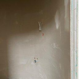 Ściana po nałożeniu tynku maszynowego, widoczne przewody elektryczne i rury kanalizacyjne przy podłodze. Świeży tynk o delikatnej fakturze.