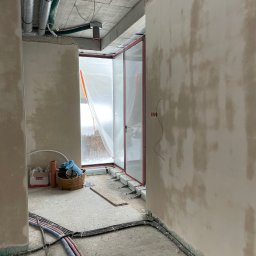 Wnętrze w trakcie prac wykończeniowych: ściany po tynkowaniu, widoczne instalacje elektryczne i rury. Okno zabezpieczone folią. Na podłodze kable i narzędzia.