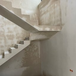Surowe schody betonowe w trakcie wykańczania gładzią, widoczne niedoskonałości i surowy beton. W tle ściany przygotowane do malowania, widoczne kable.