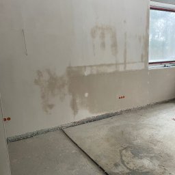 Ściana po nałożeniu gładzi, widoczne ślady wilgoci i niedoskonałości przed szlifowaniem. Na podłodze widoczne ślady po instalacjach. Surowy stan wykończenia wnętrza.