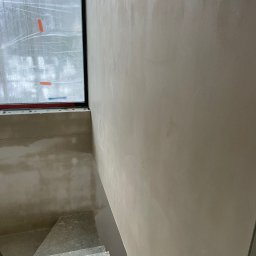 Portal-tech - Świeżo nałożona, gładka, jasna gładź na ścianie klatki schodowej, widoczna z góry. Okno z folią ochronną w tle. Surowy beton schodów.