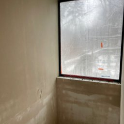 Portal-tech - Ściana po nałożeniu gładzi, z widocznymi śladami szpachlowania. Okno zabezpieczone folią. Surowy betonowy sufit. Wystające kable elektryczne.