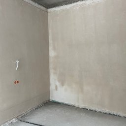 Portal-tech - Wnętrze pokoju po nałożeniu gładzi, widoczne ściany w jasnym odcieniu beżu, surowy betonowy sufit i przygotowana podłoga. Na ścianie widoczne gniazdka elektryczne.