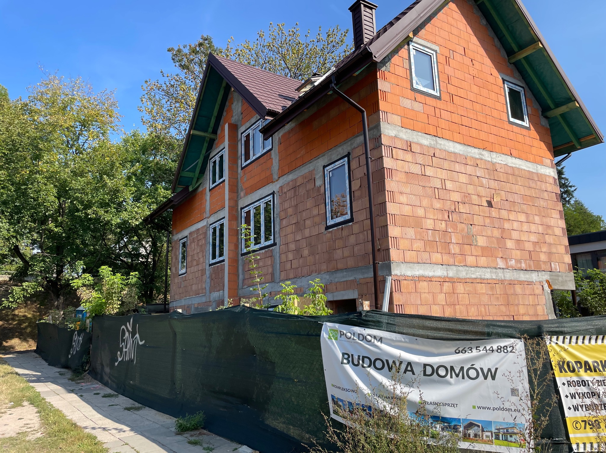 Dwukondygnacyjny dom w trakcie budowy, z widoczną konstrukcją z cegieł i zamontowanymi oknami. Na pierwszym planie baner reklamowy firmy budowlanej.
