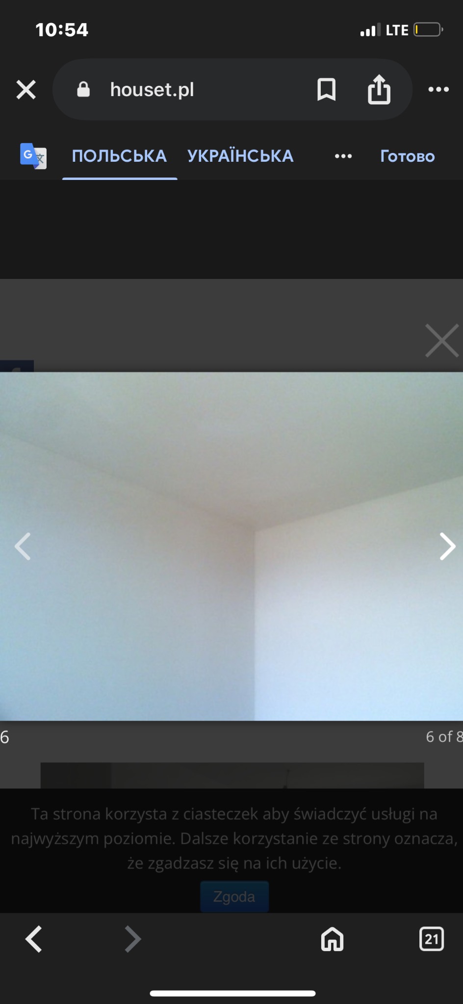 Puste, pomalowane na biało wnętrze pokoju z narożnikiem, widziane przez ekran telefonu. Widoczne elementy interfejsu strony internetowej.