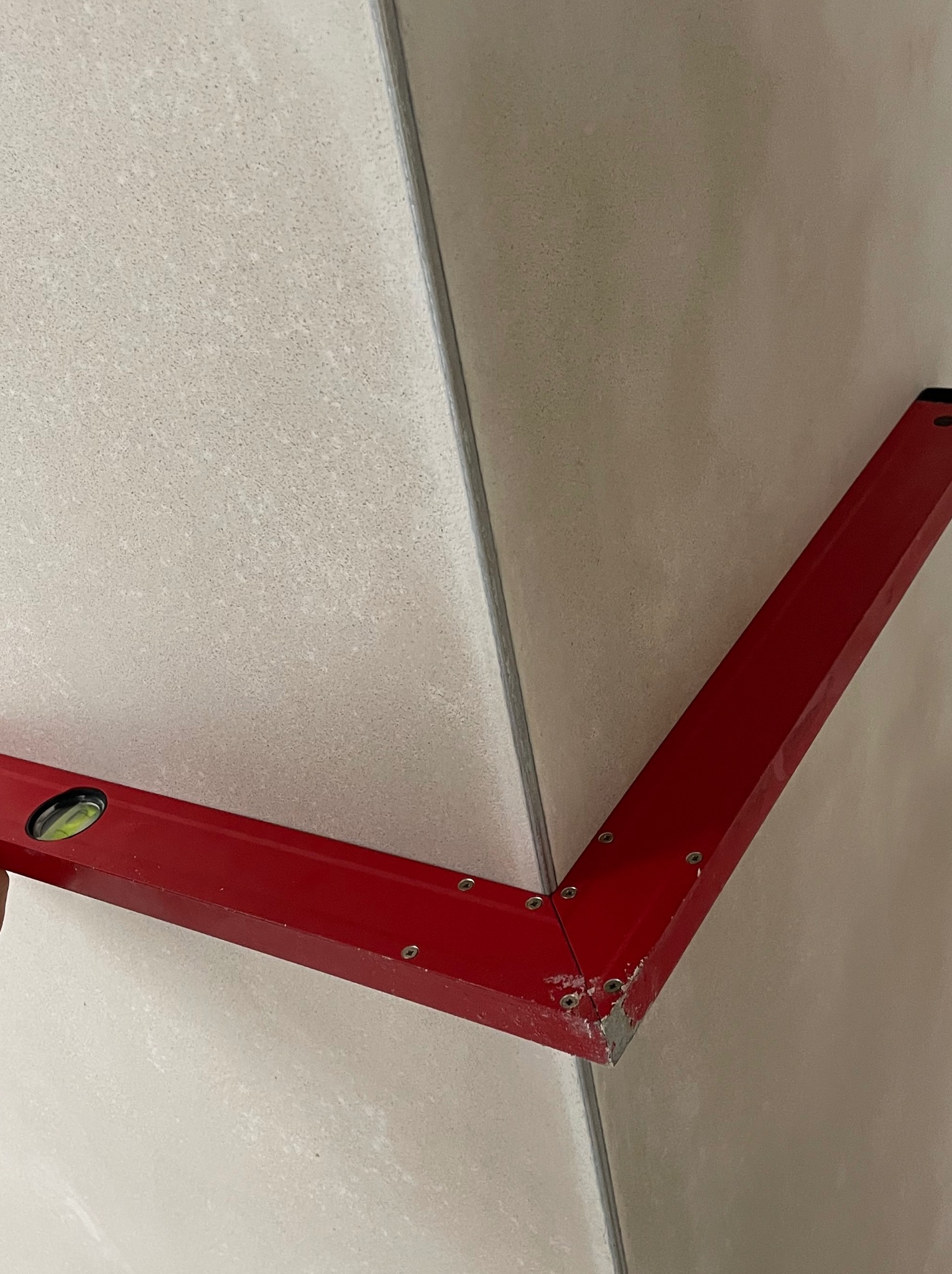 Poziomica kątowa na jasnych płytkach ściennych, sprawdzanie kąta prostego w rogu pomieszczenia. Widoczne ślady zaprawy na narzędziu.