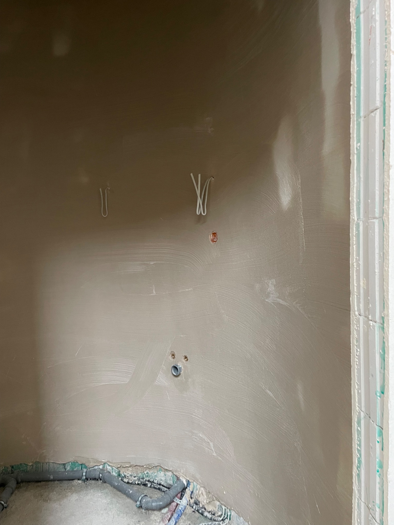 Ściana po nałożeniu tynku maszynowego, widoczne przewody elektryczne i rury kanalizacyjne przy podłodze. Świeży tynk o delikatnej fakturze.