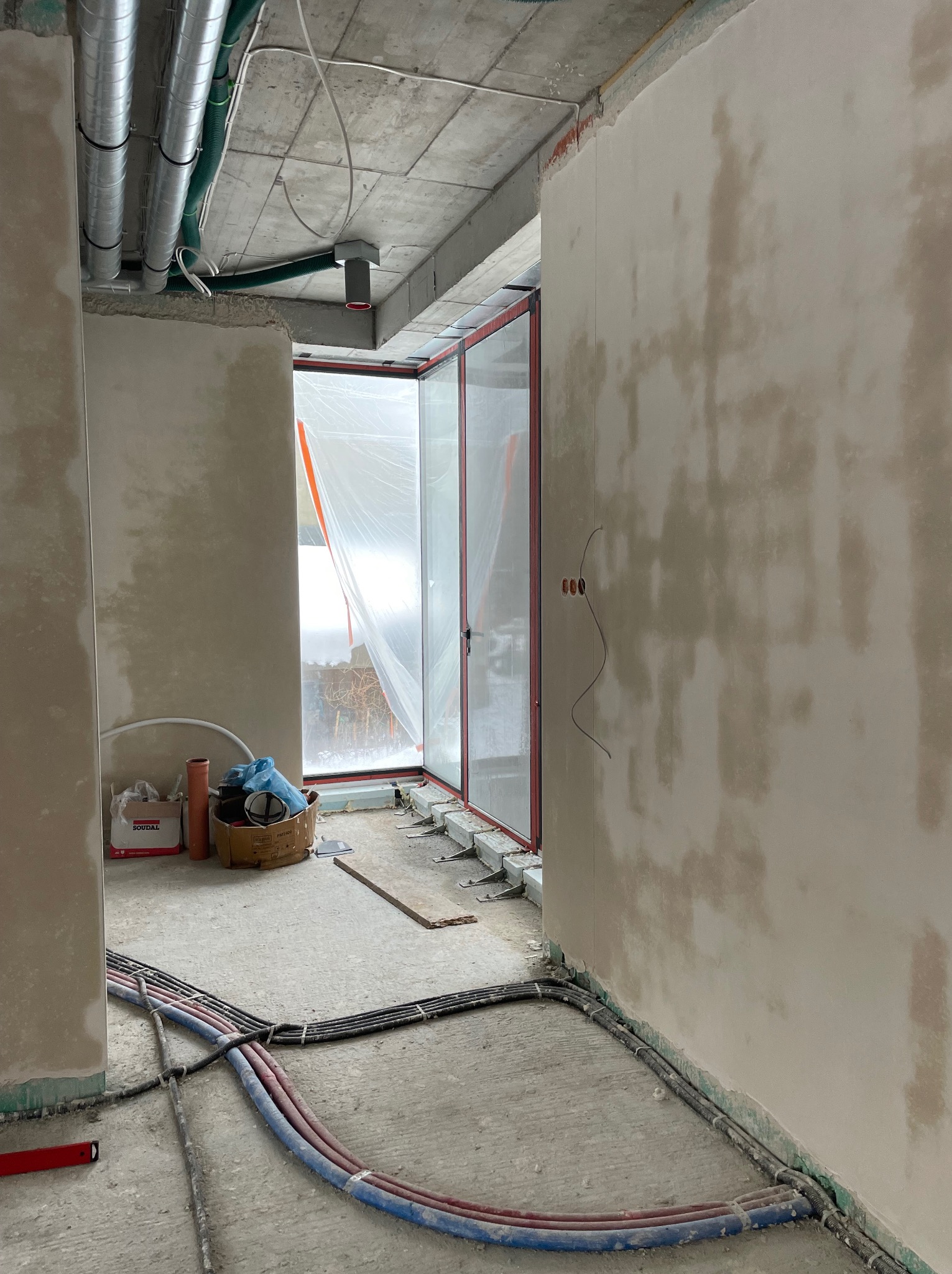 Wnętrze w trakcie prac wykończeniowych: ściany po tynkowaniu, widoczne instalacje elektryczne i rury. Okno zabezpieczone folią. Na podłodze kable i narzędzia.