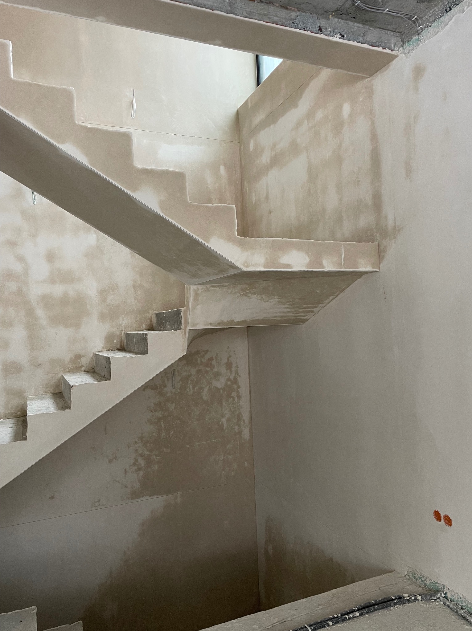 Surowe schody betonowe w trakcie wykańczania gładzią, widoczne niedoskonałości i surowy beton. W tle ściany przygotowane do malowania, widoczne kable.