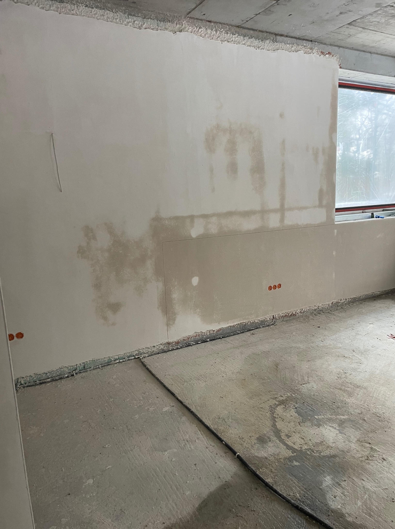 Ściana po nałożeniu gładzi, widoczne ślady wilgoci i niedoskonałości przed szlifowaniem. Na podłodze widoczne ślady po instalacjach. Surowy stan wykończenia wnętrza.