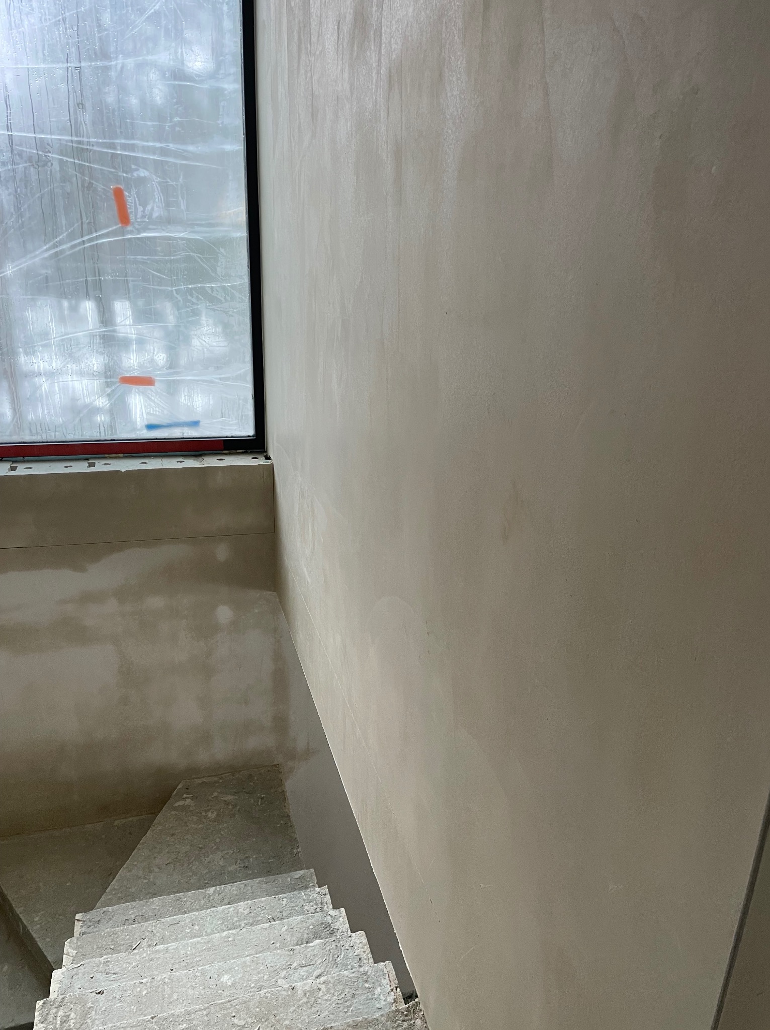 Świeżo nałożona, gładka, jasna gładź na ścianie klatki schodowej, widoczna z góry. Okno z folią ochronną w tle. Surowy beton schodów.
