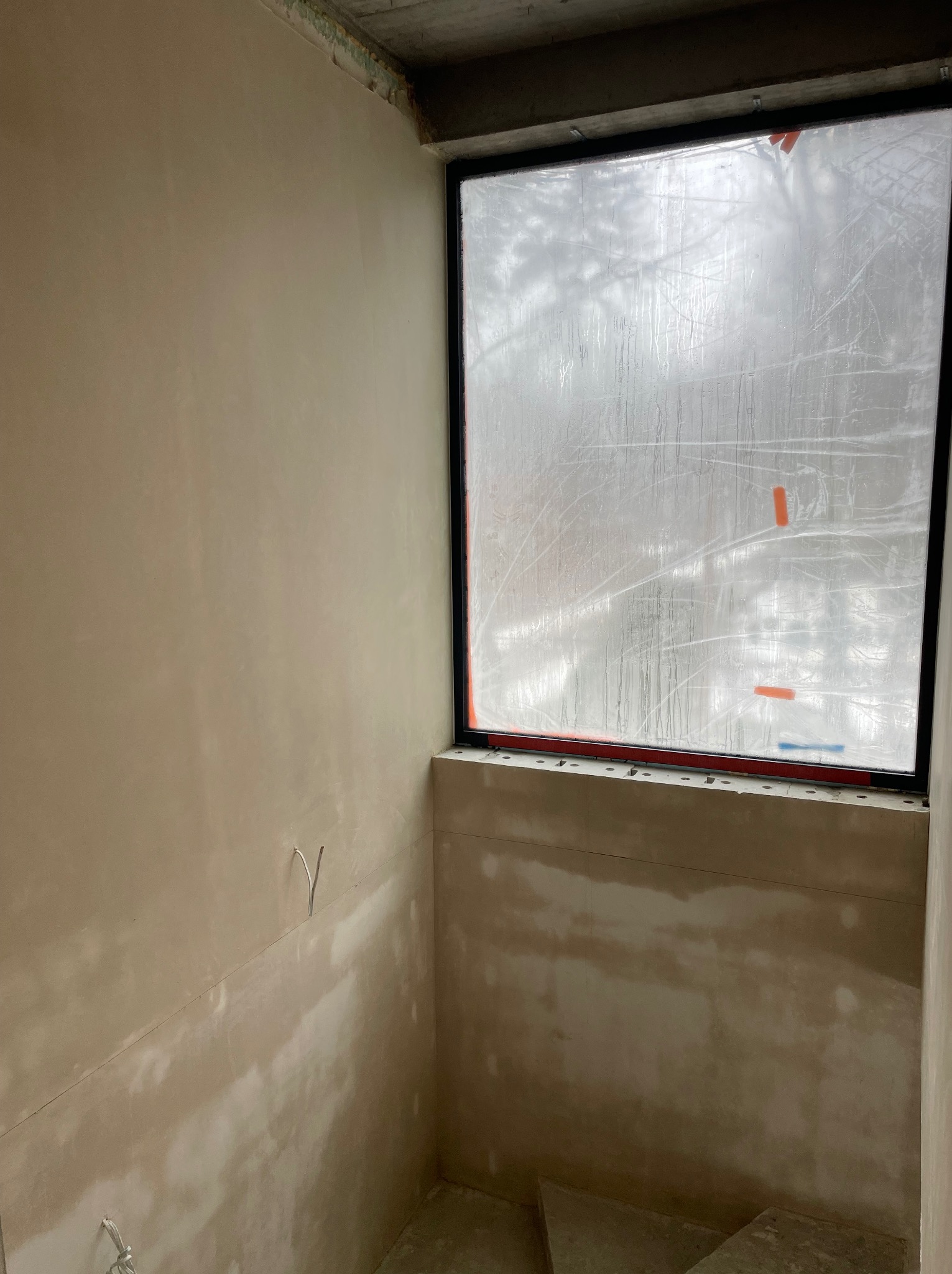 Ściana po nałożeniu gładzi, z widocznymi śladami szpachlowania. Okno zabezpieczone folią. Surowy betonowy sufit. Wystające kable elektryczne.