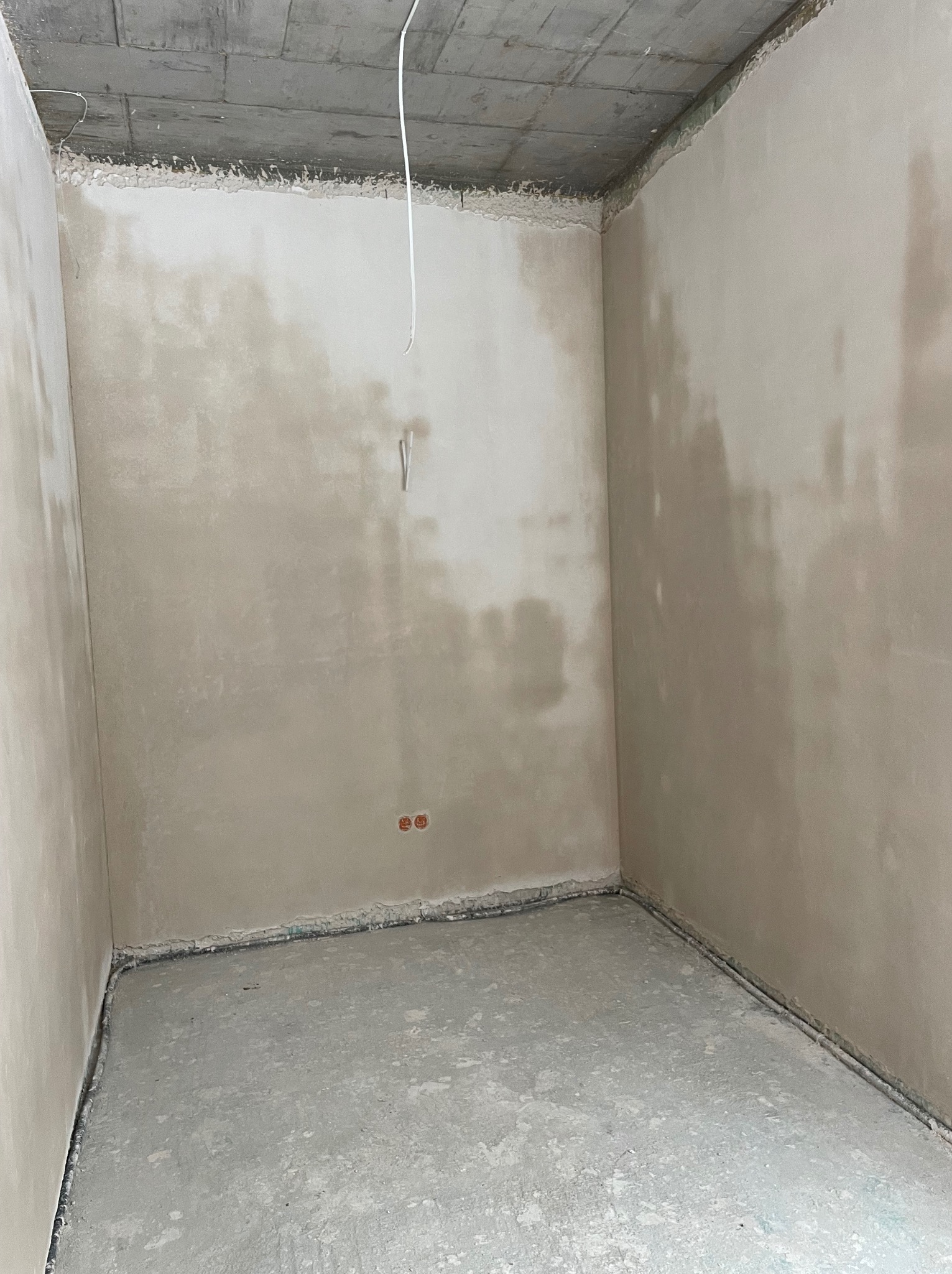 Surowe wnętrze pokoju po tynkowaniu ścian i wylewce podłogi. Widoczne wystające kable i surowy betonowy strop. Ściany w odcieniach beżu.