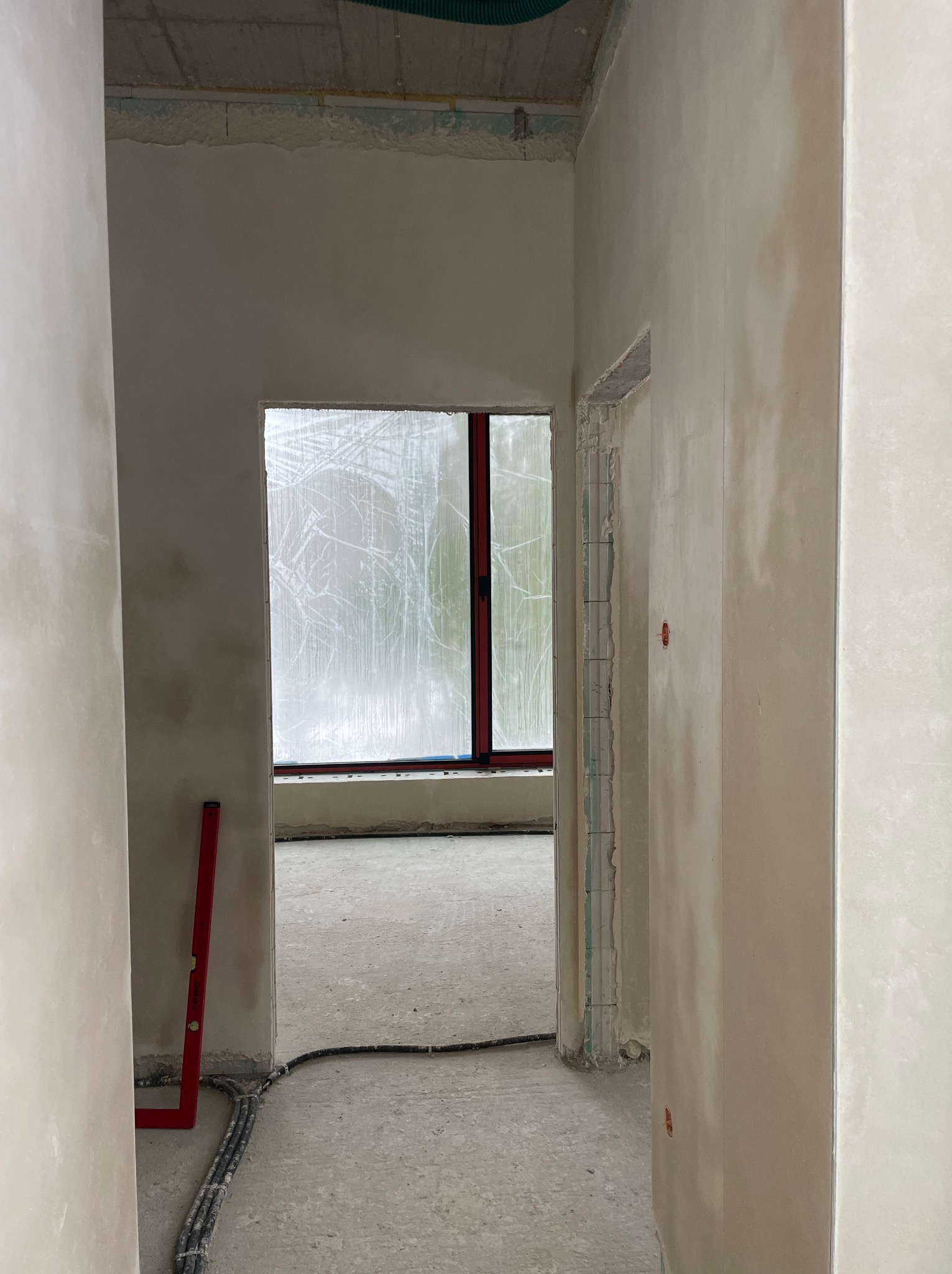 Wnętrze w trakcie prac wykończeniowych: ściany po tynkowaniu, widoczne przewody elektryczne na podłodze, poziomica oparta o ścianę. Okno zabezpieczone folią.