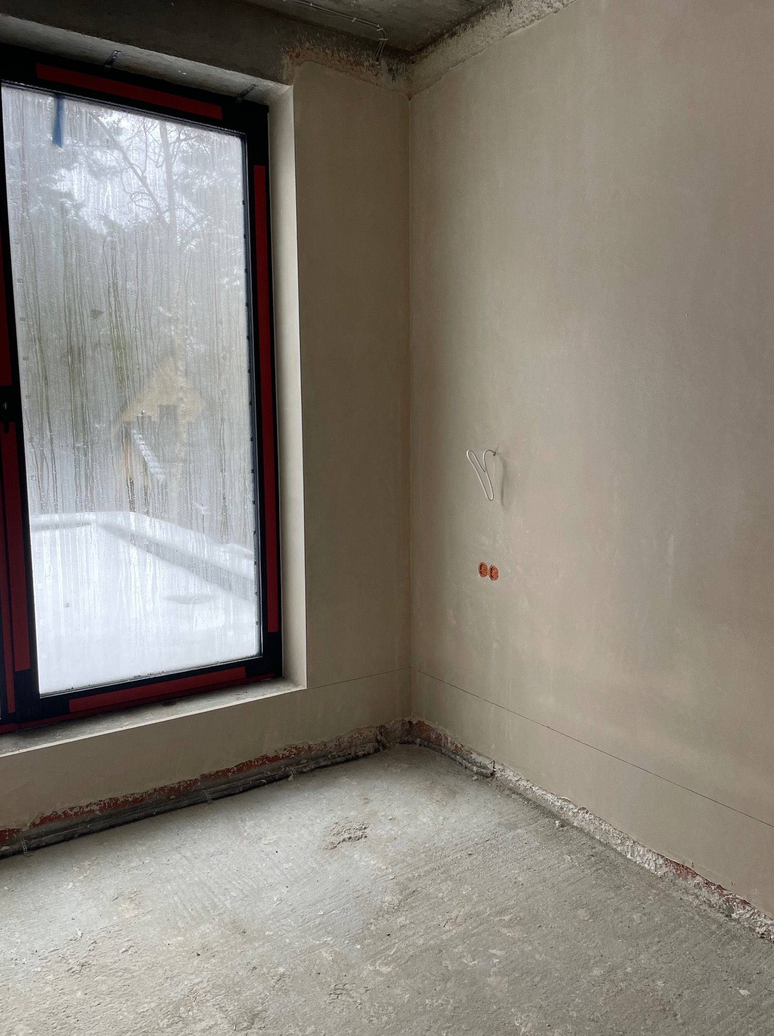 Pokój po tynkowaniu maszynowym: gładkie ściany, okno z widokiem na zimowy krajobraz, widoczne instalacje elektryczne. Surowy beton na podłodze.