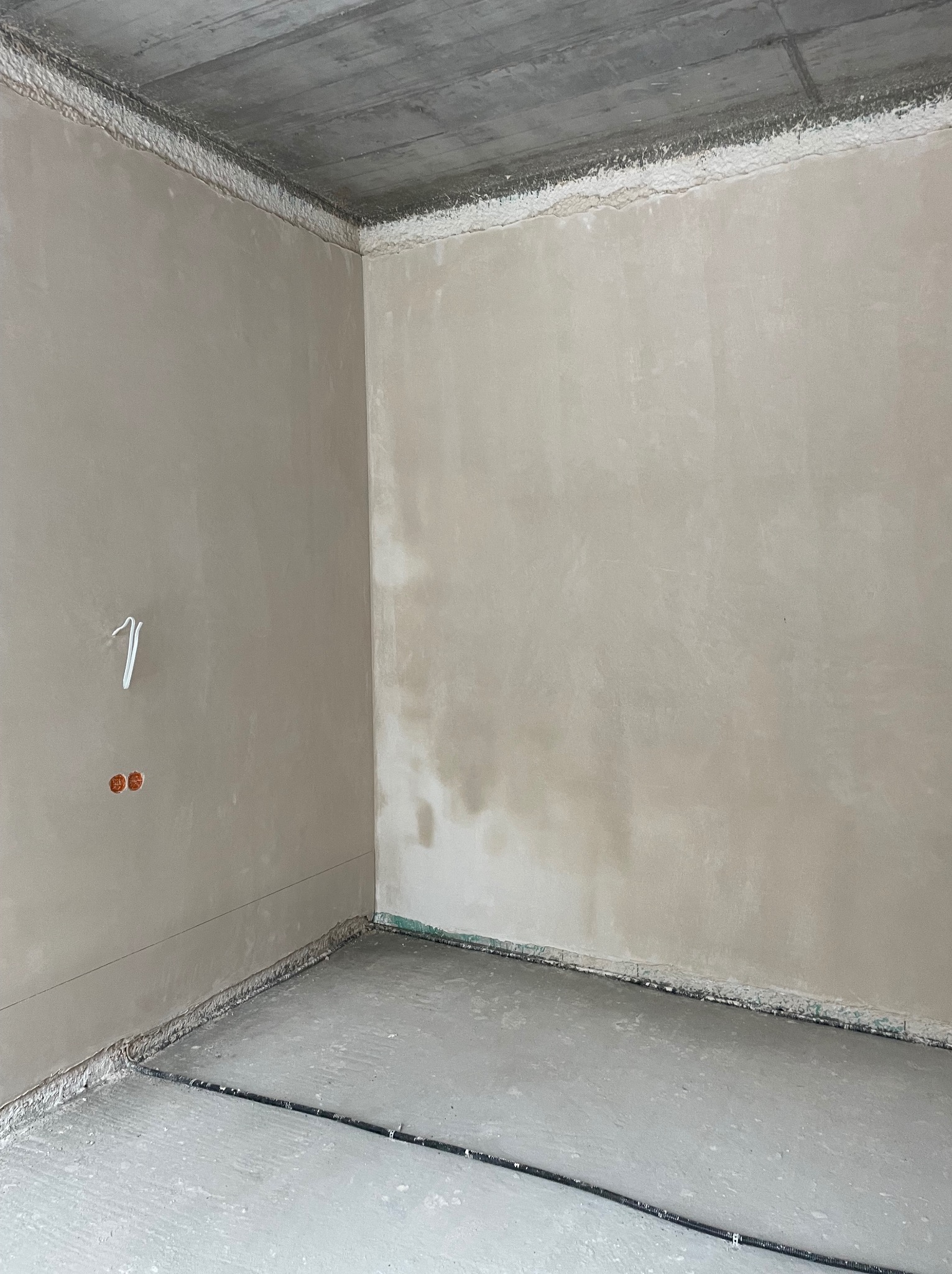 Wnętrze pokoju po nałożeniu gładzi, widoczne ściany w jasnym odcieniu beżu, surowy betonowy sufit i przygotowana podłoga. Na ścianie widoczne gniazdka elektryczne.