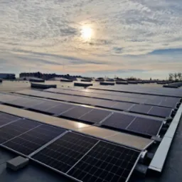 Widok z góry na dużą instalację fotowoltaiczną na dachu przemysłowym, z widocznym niebem z chmurami i słońcem odbijającym się w panelach.