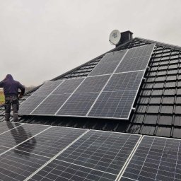 FOTON-SERWIS USŁUGI OGÓLNOBUDOWLANE SEBASTIAN KUC - Instalator w ciemnej bluzie montuje panele fotowoltaiczne na czarnym dachu z dachówki ceramicznej, widoczna antena satelitarna i pochmurne niebo w tle.