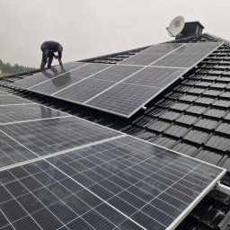 FOTON-SERWIS USŁUGI OGÓLNOBUDOWLANE SEBASTIAN KUC - Montaż paneli słonecznych na dachu z czarnej dachówki, widoczna sylwetka instalatora w odzieży roboczej i pomarańczowych rękawicach, w tle antena satelitarna i pochmurne niebo.