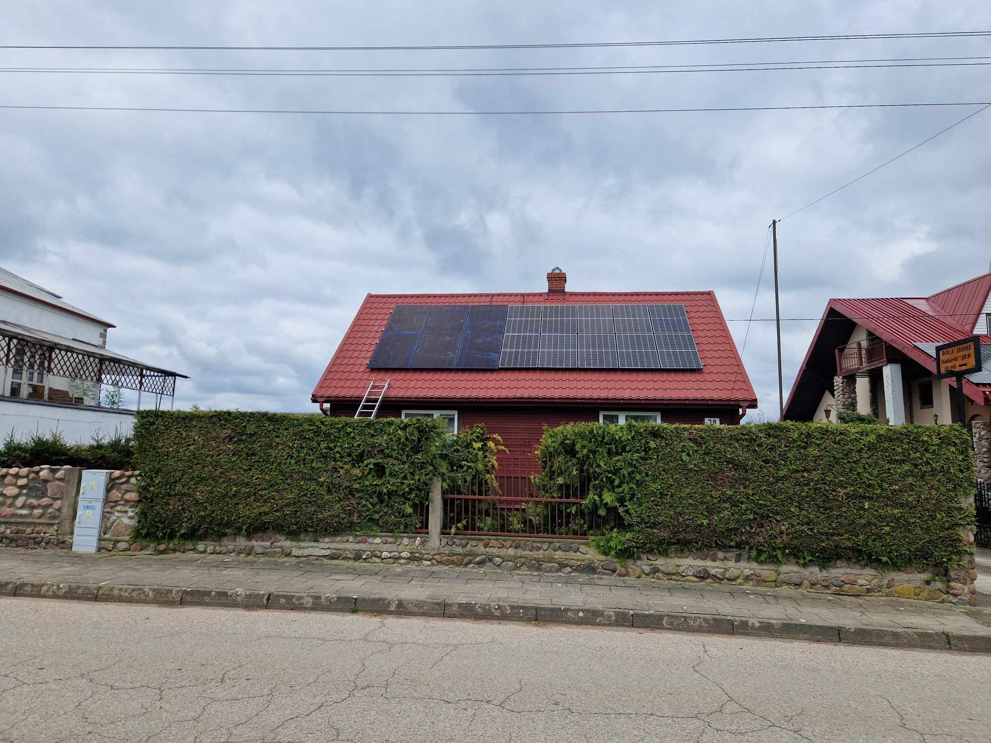 Czerwony dom jednorodzinny z panelami słonecznymi na dachu, drabina oparta o dach, otoczony zielonym żywopłotem i kamiennym murkiem.