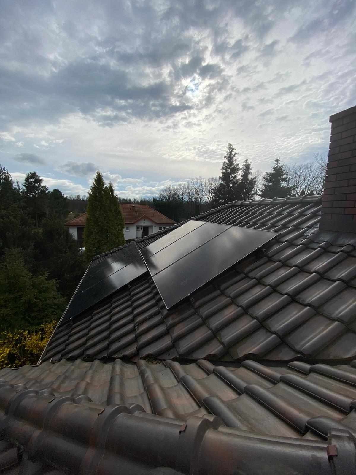 Ciemne panele słoneczne zamontowane na brązowym, ceramicznym dachu domu w Białymstoku, widok z góry na tle zachmurzonego nieba.