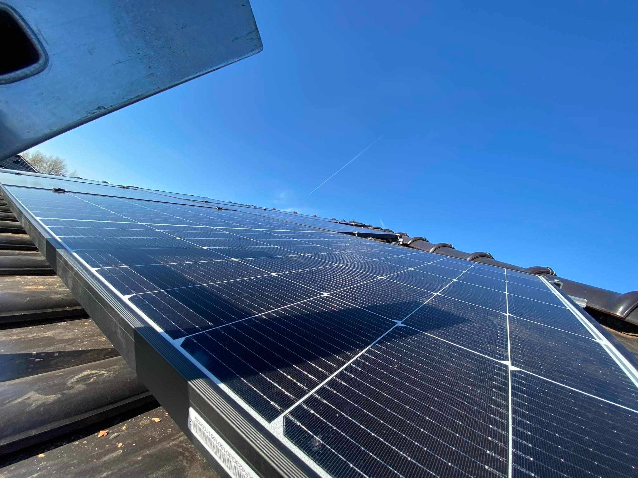 Panel słoneczny na dachu z dachówki ceramicznej, widok z dołu pod kątem, błękitne niebo w tle, ślad po samolocie.