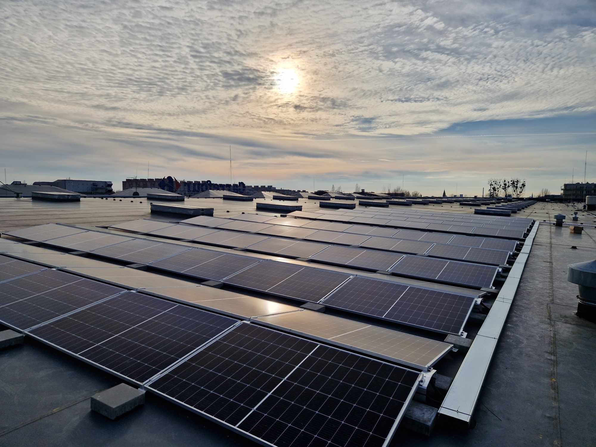 Widok z góry na dużą instalację fotowoltaiczną na dachu przemysłowym, z widocznym niebem z chmurami i słońcem odbijającym się w panelach.