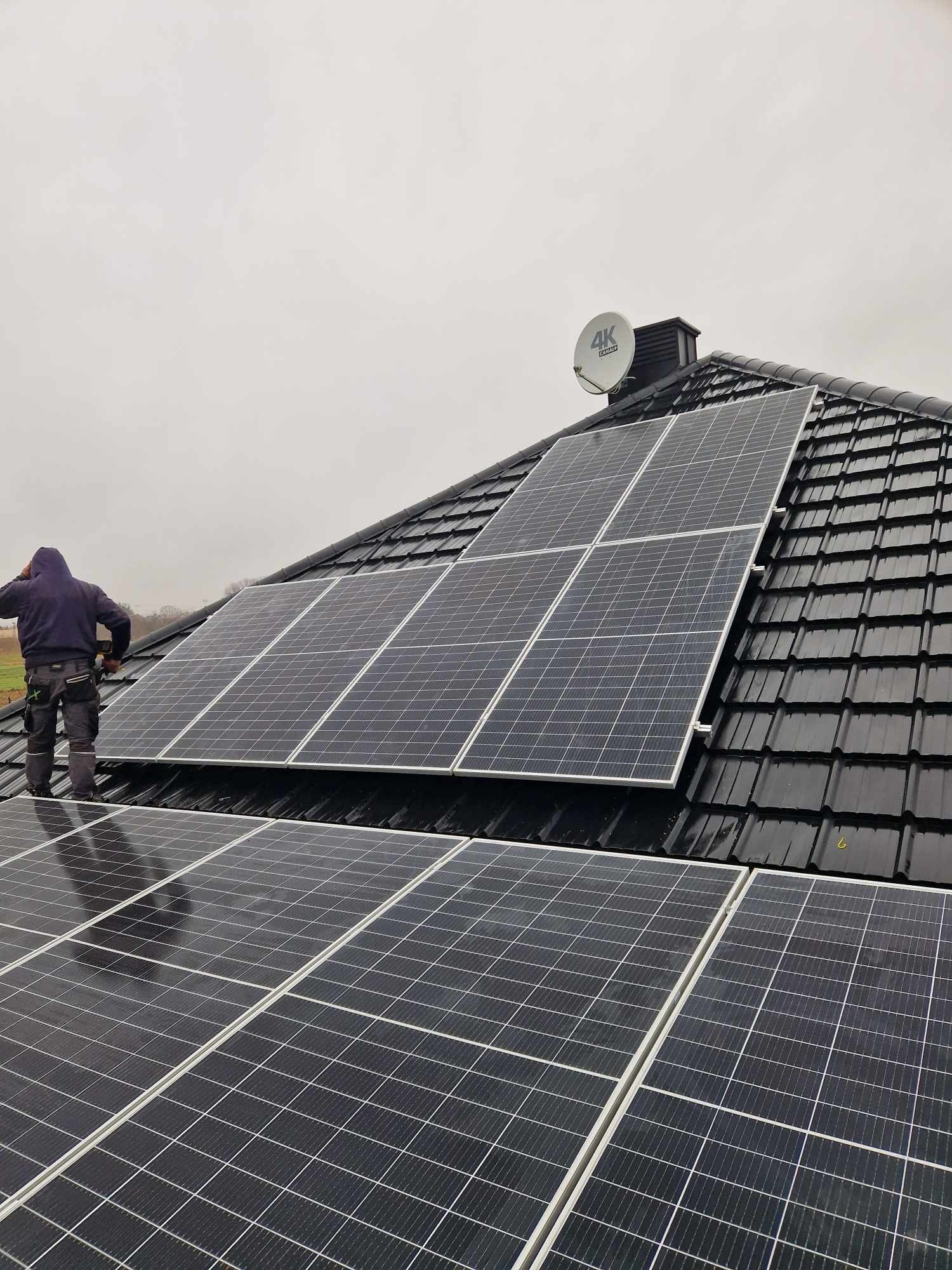Instalator w ciemnej bluzie montuje panele fotowoltaiczne na czarnym dachu z dachówki ceramicznej, widoczna antena satelitarna i pochmurne niebo w tle.