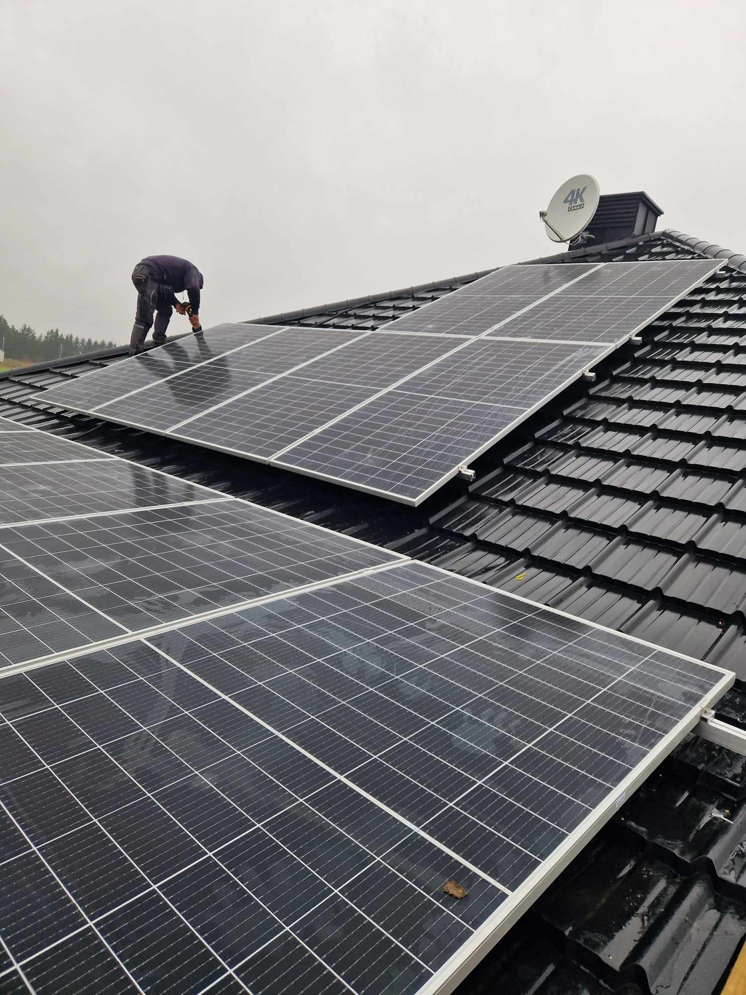 Montaż paneli słonecznych na dachu z czarnej dachówki, widoczna sylwetka instalatora w odzieży roboczej i pomarańczowych rękawicach, w tle antena satelitarna i pochmurne niebo.