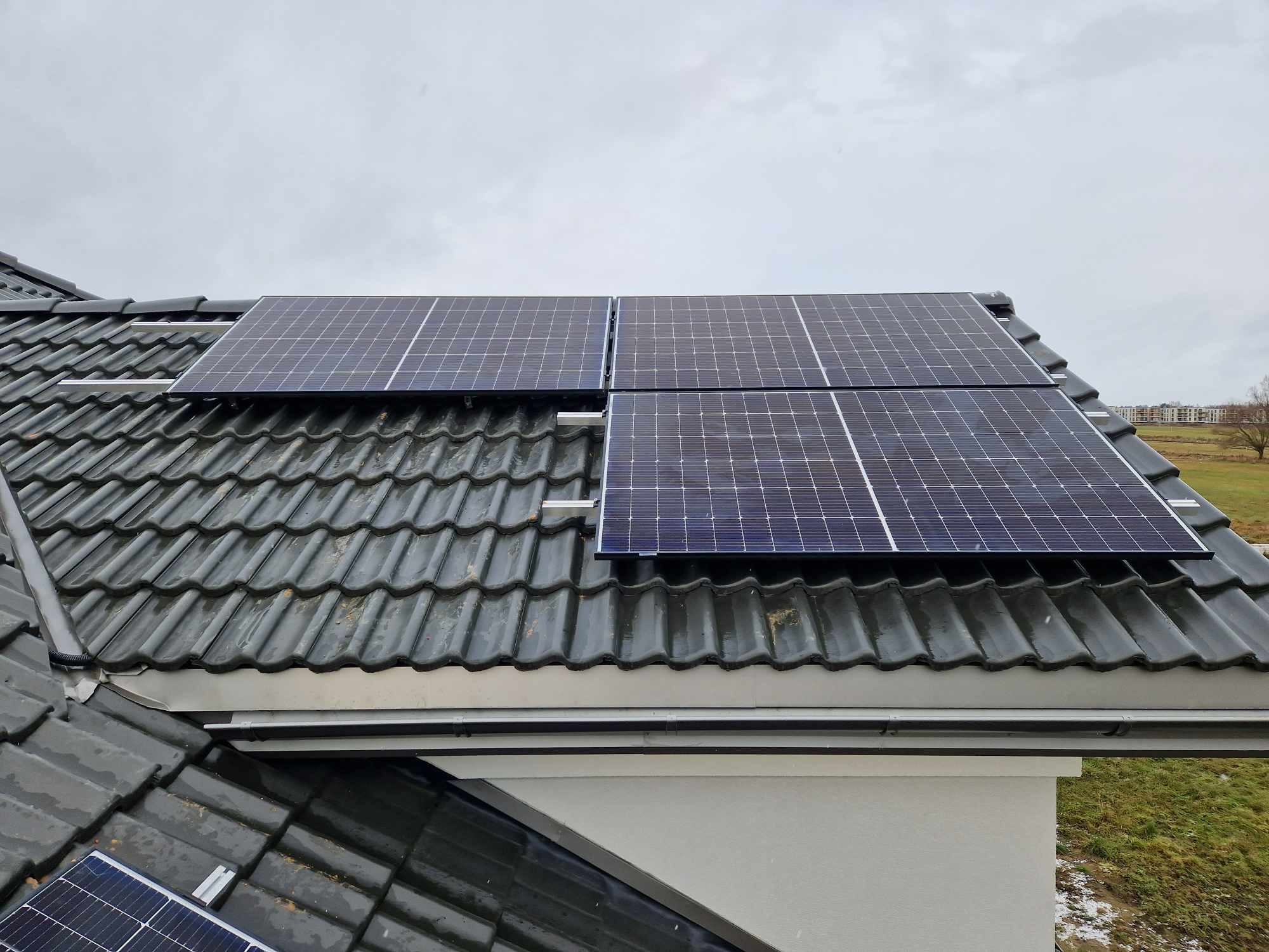 Panele słoneczne zamontowane na ciemnym, błyszczącym dachu z dachówki ceramicznej, widok z góry na tle pochmurnego nieba.