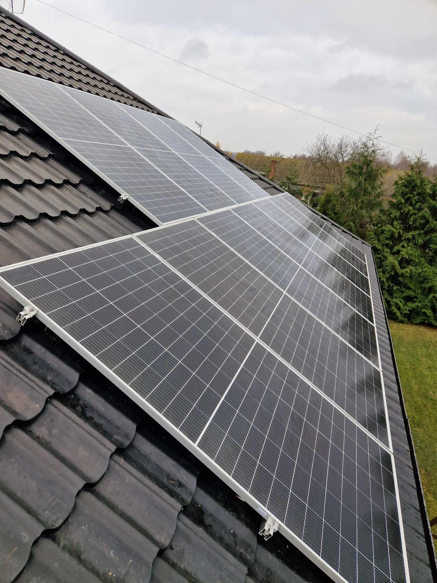 Panele słoneczne zamontowane na ciemnej, falistej dachówce, widoczne mocowania paneli i fragment zielonego trawnika w tle.