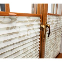 ZAKŁAD USŁUGOWO-HANDLOWY JULITA JURKIEWICZ - Zamknięte, kremowe, plisowane osłony okienne z delikatnym, roślinnym wzorem, zamontowane w brązowych, drewnianych oknach z mosiężnymi klamkami.