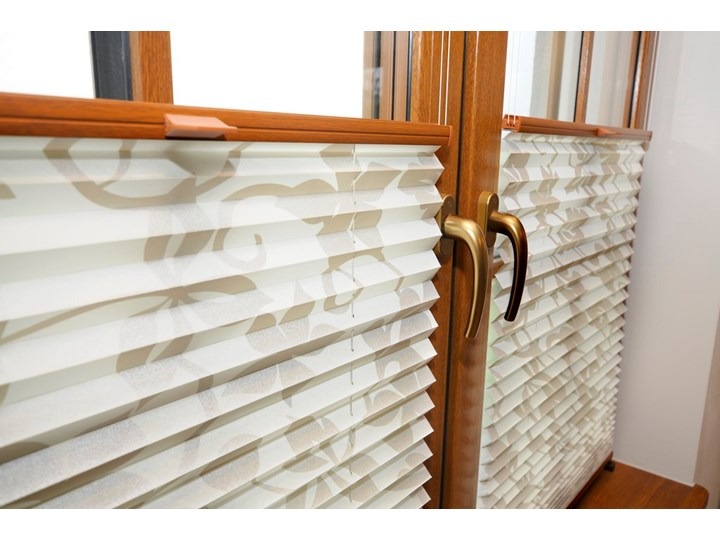 Zamknięte, kremowe, plisowane osłony okienne z delikatnym, roślinnym wzorem, zamontowane w brązowych, drewnianych oknach z mosiężnymi klamkami.