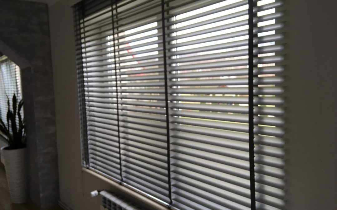 Szare poziome lamele okienne z widocznym kaloryferem pod oknem i rośliną doniczkową w tle.