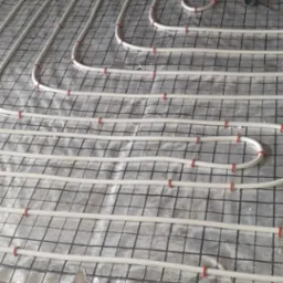 Białe rury ogrzewania podłogowego zamocowane czerwonymi klipsami na czarnej siatce, ułożone na srebrnej folii izolacyjnej, z niebieskim agregatem chłodniczym w tle.