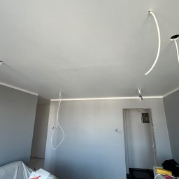 Remonty DL Dominik Lipiec - Szare wnętrze w trakcie remontu: widoczne świeżo pomalowane ściany, wystające przewody elektryczne z sufitu i zabezpieczone meble folią malarską.