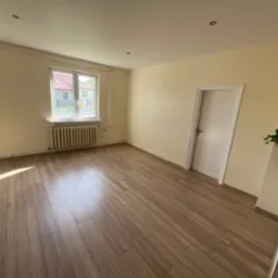 Jasne wnętrze po remoncie: świeżo ułożone panele podłogowe w odcieniach drewna, kremowe ściany, białe drzwi i okno z widokiem na zieleń. Grzejnik pod oknem.