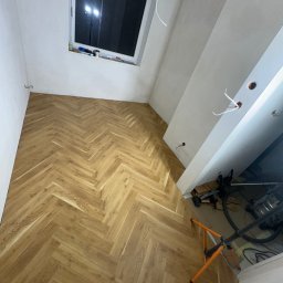 Remonty DL Dominik Lipiec - Ukończona podłoga z paneli ułożonych w jodełkę w jasnym pomieszczeniu z oknem. Widoczne narzędzia pracy i lampa oświetleniowa.
