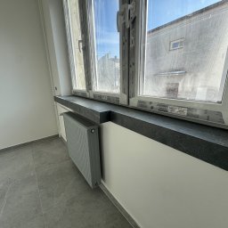 Remonty DL Dominik Lipiec - Wnętrze po remoncie: szare płytki podłogowe, grzejnik w kolorze ścian, okno PCV z czarnym parapetem. Widok na budynek przez okno.