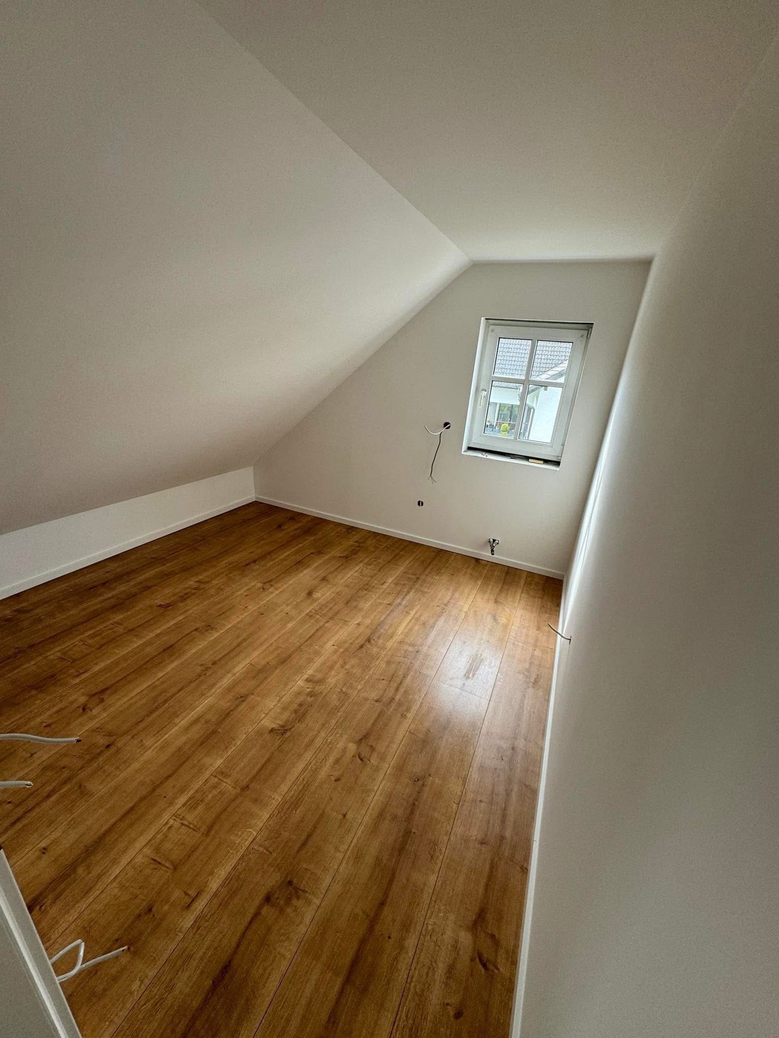 Pokój na poddaszu po remoncie: świeżo położone panele podłogowe w odcieniach brązu, białe ściany z widocznymi przewodami elektrycznymi i małe okno dachowe z widokiem na zewnątrz.