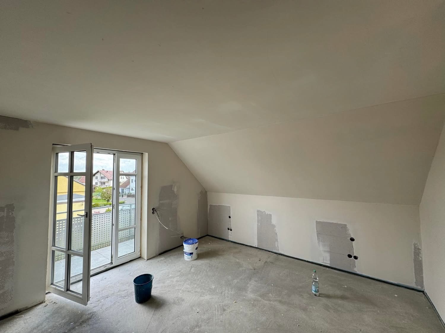 Pokój na poddaszu w trakcie remontu: ściany z miejscowymi łatami gładzi, otwarte drzwi balkonowe, wiadro z farbą, niebieskie wiadro i butelka wody na podłodze.