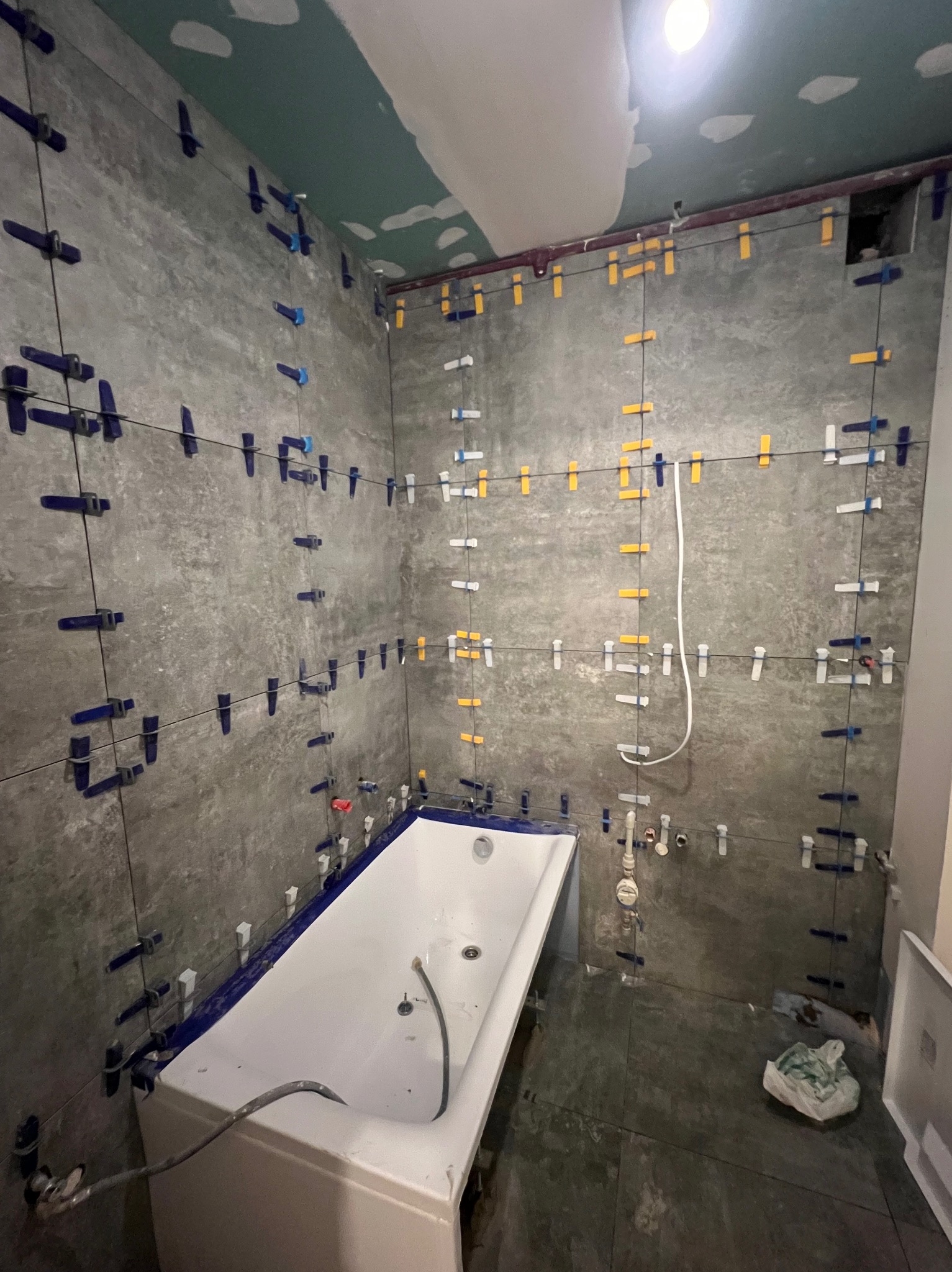 Łazienka w trakcie remontu, widoczne szare płytki na ścianach z systemem poziomowania, biała wanna zabezpieczona folią, sufit w kolorze zielonym, widoczne instalacje hydrauliczne.