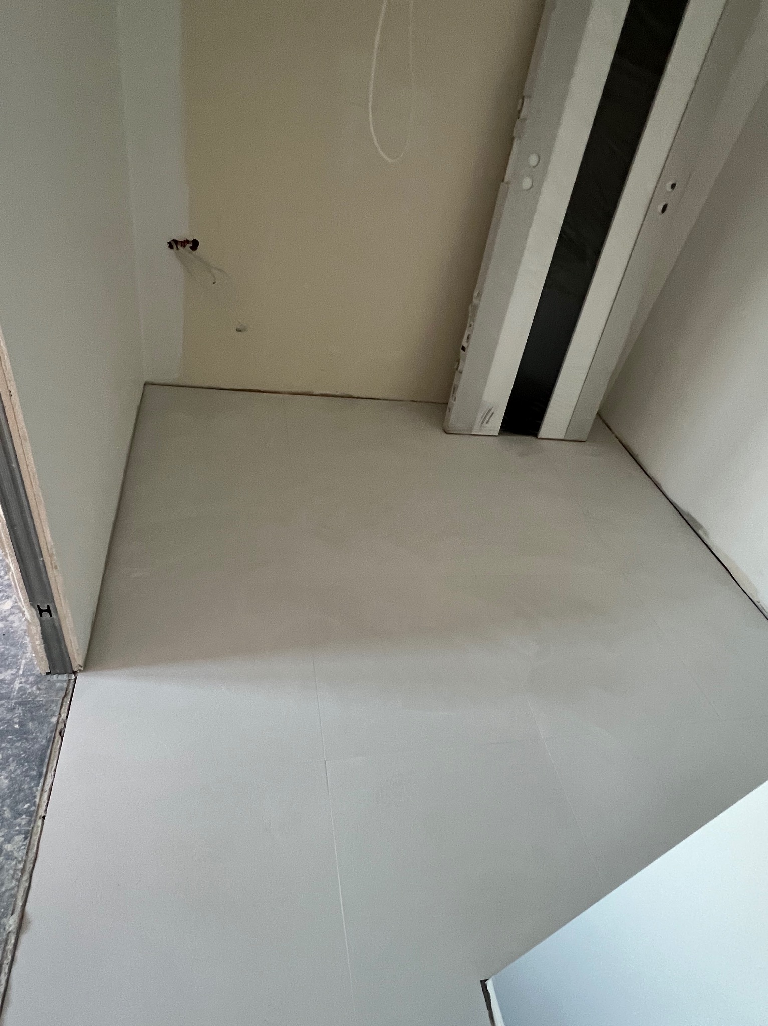 Ułożone jasnoszare płytki podłogowe w pomieszczeniu w trakcie remontu, widoczne fragmenty ścian w różnych odcieniach bieli i beżu, oraz wystające kable elektryczne.