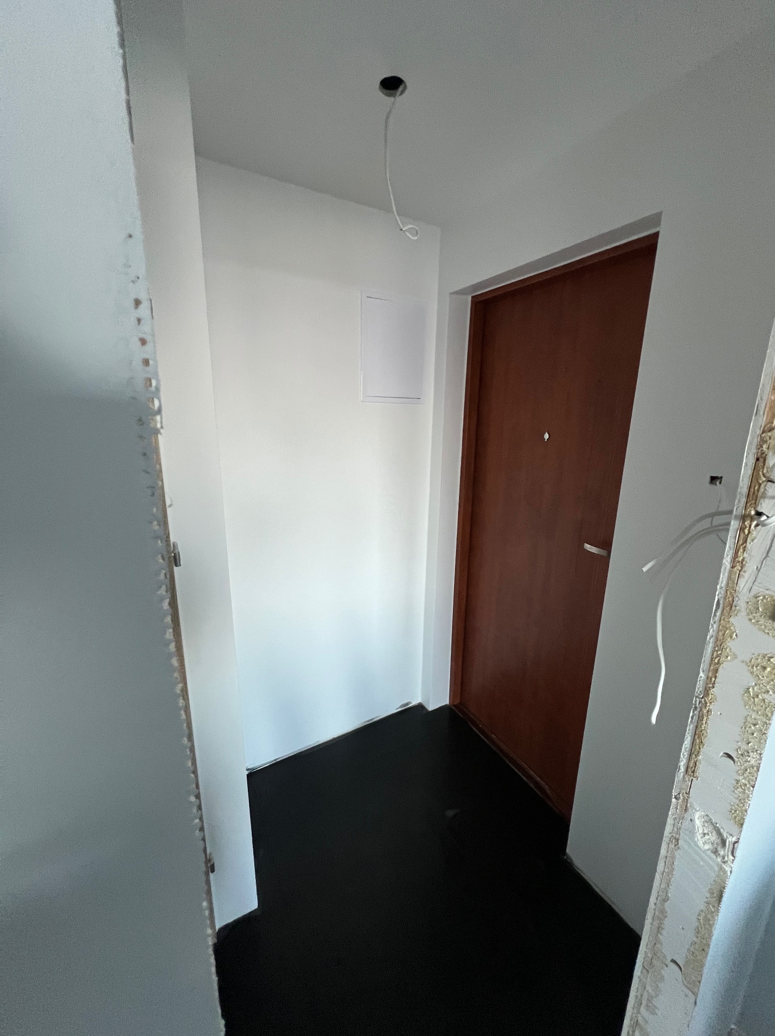 Korytarz w trakcie remontu: świeżo pomalowane ściany na biało, ciemna podłoga, brązowe drzwi z metalową klamką, widoczne kable wystające z sufitu i fragmenty starej futryny.